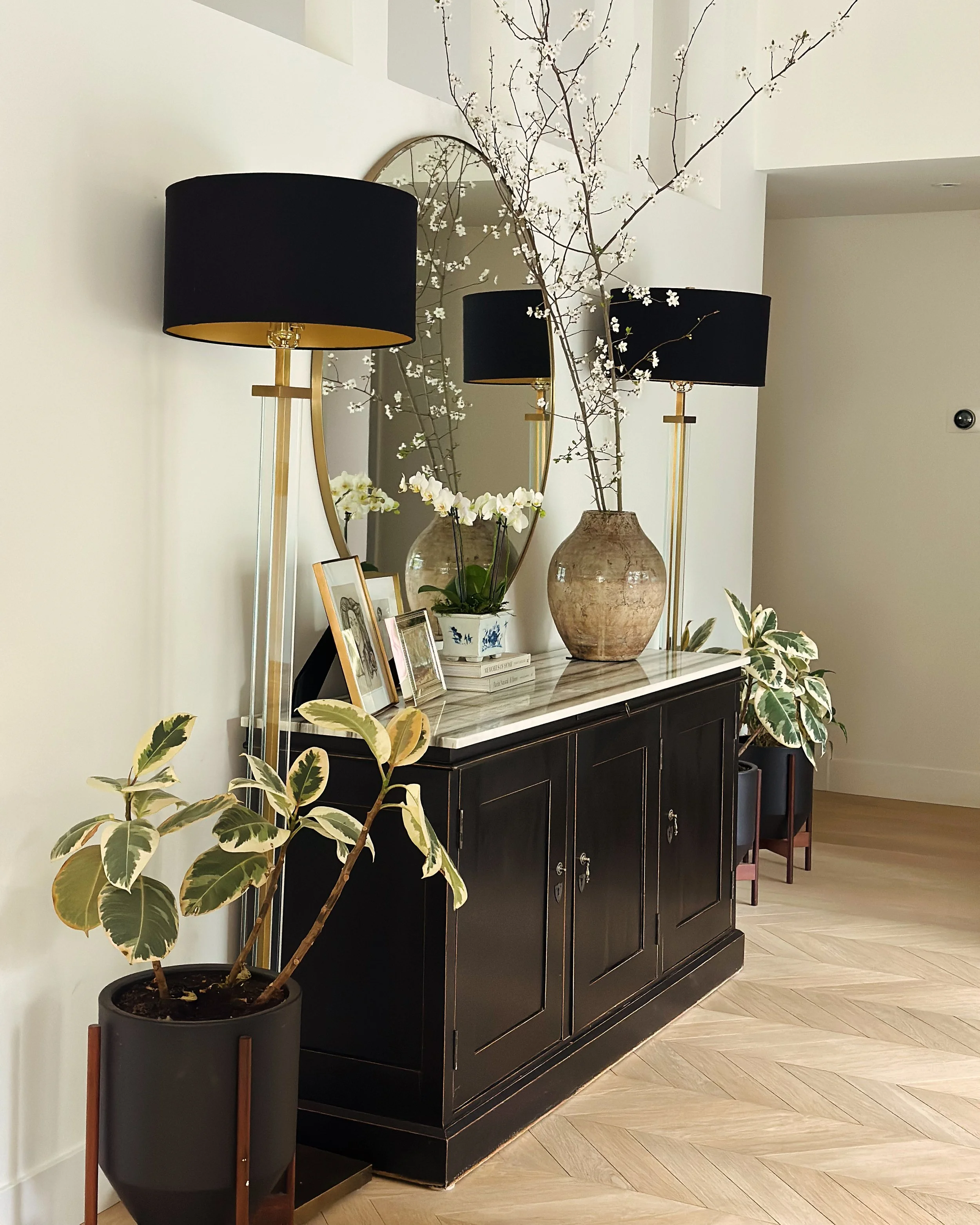 A black sideboard with a marble top, decorated with photo frames, a large ceramic vase with white flowers and branches, a round mirror, two black lamps with gold bases, and potted plants serving as accents.