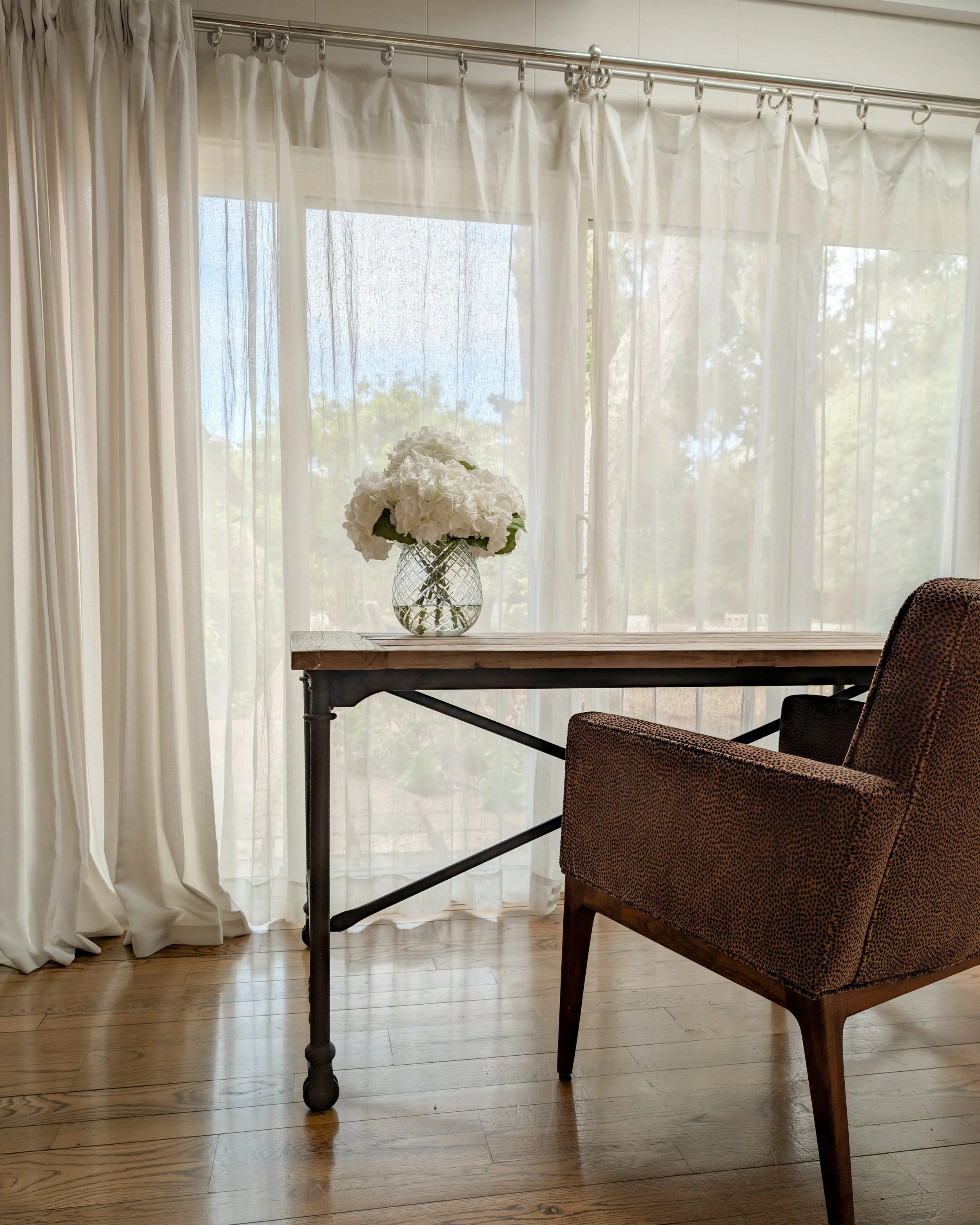 A living room corner with a wooden table, a brown upholstered chair, and a vase of white flowers on the table. Behind is a large window with sheer white curtains.
