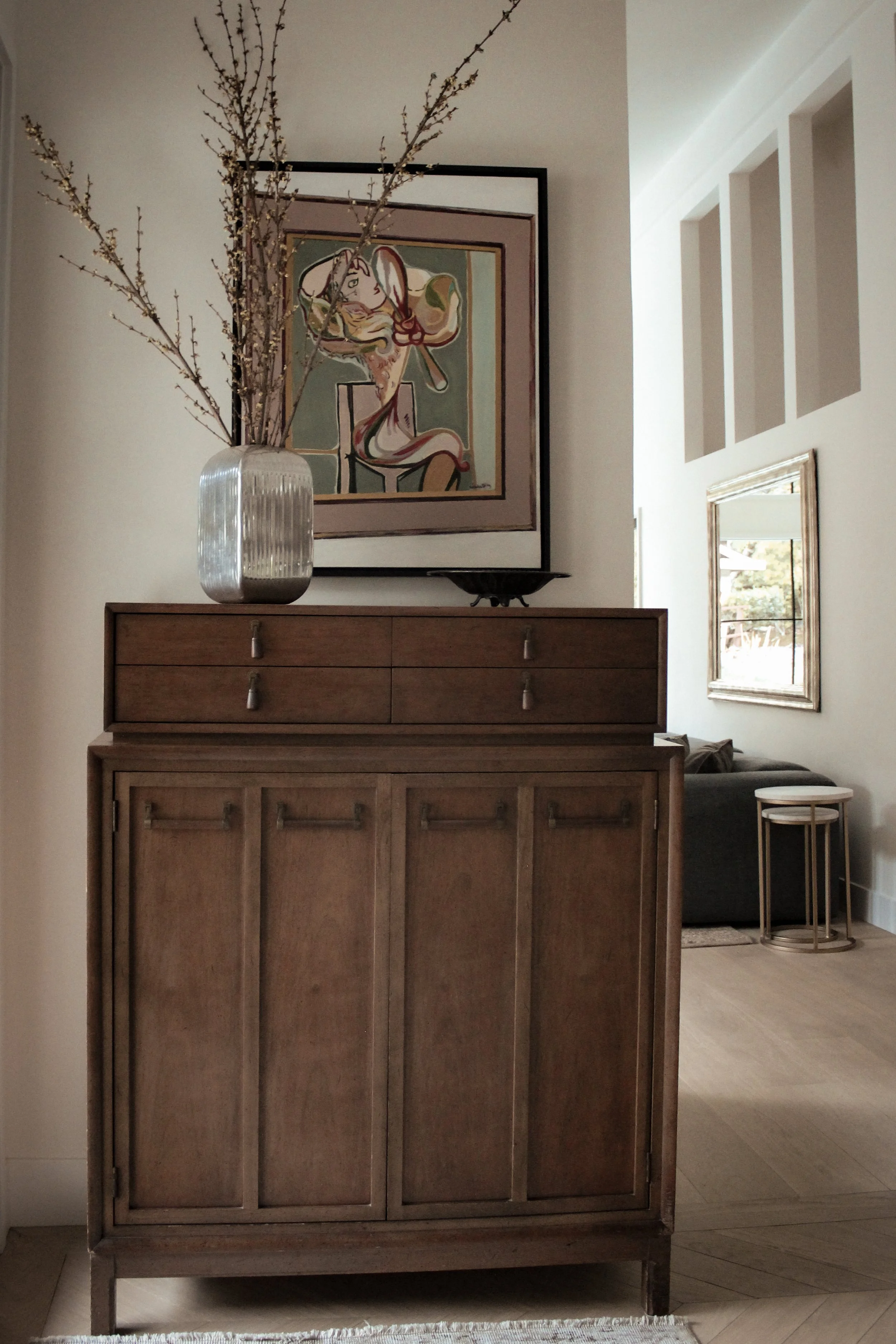 A wooden sideboard with a large vase of branches on top, a modern framed artwork behind the vase, and a small black tray. In the background, a living room with a sofa, side tables, a mirror on the wall, and natural light coming through the window.
