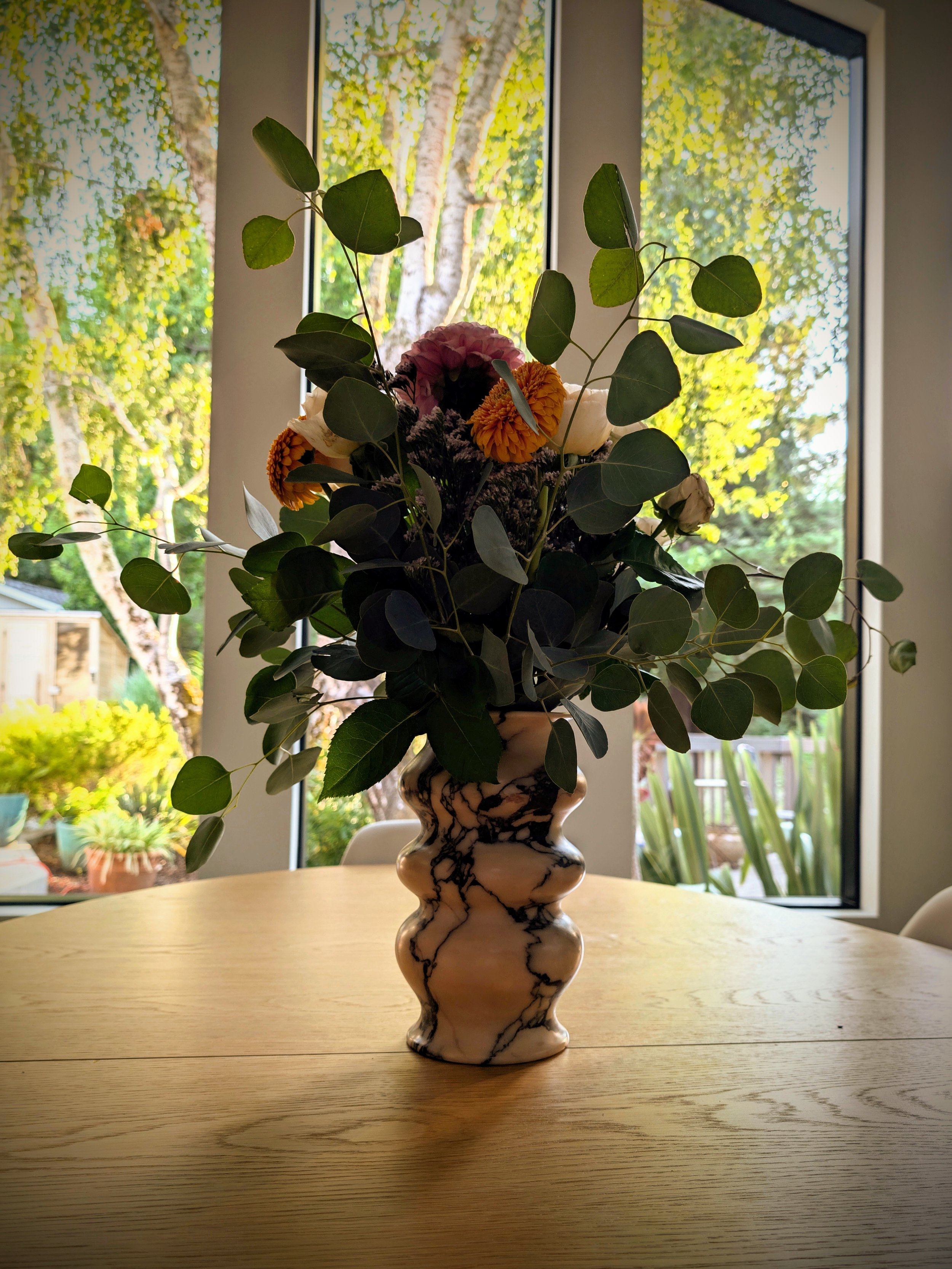 A bouquet of flowers in a marbled vase placed on a wooden table near large windows with a view of trees and a garden outside.