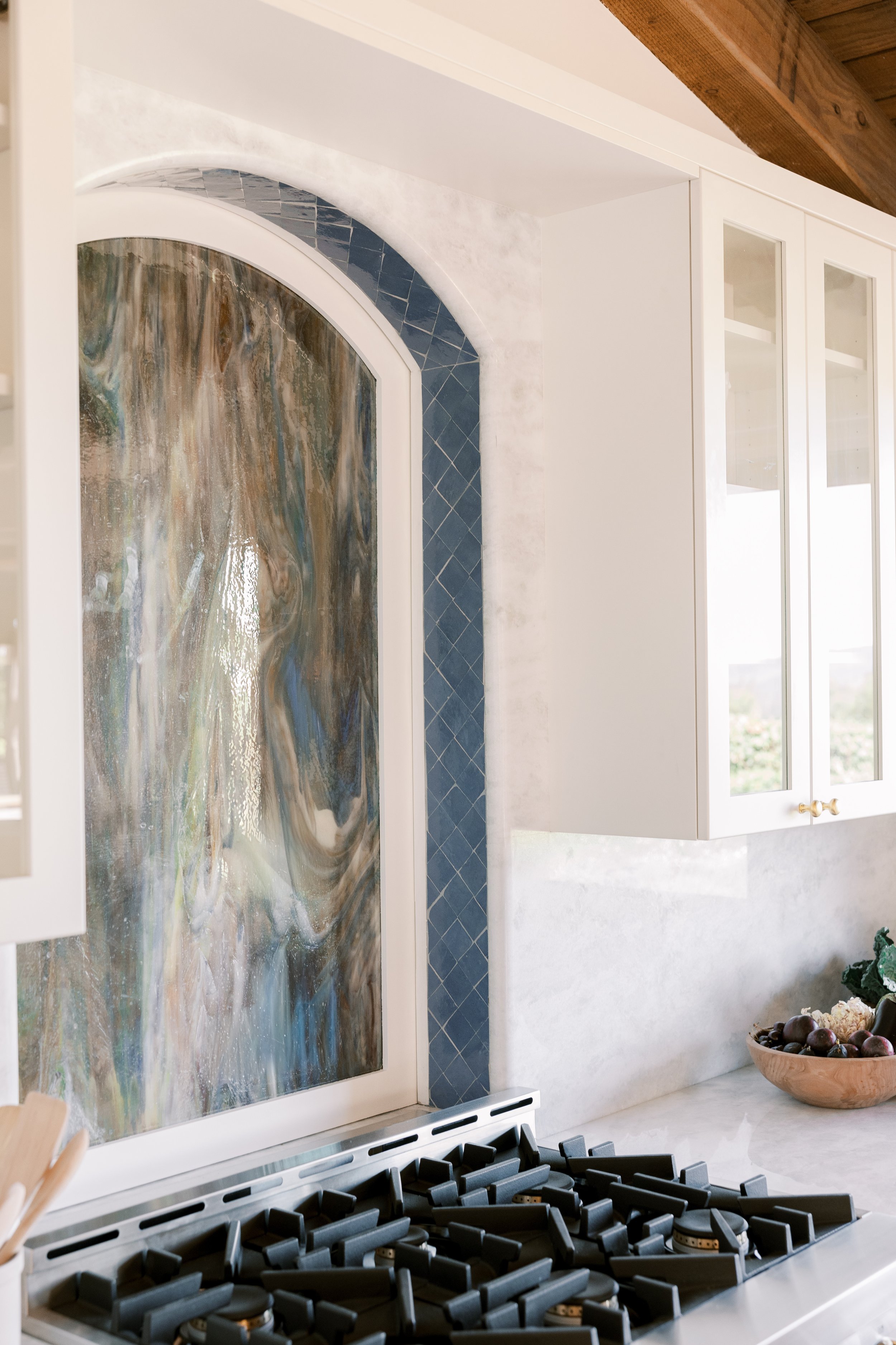 Kitchen with a stove, a glass-front cabinet, a bowl of grapes, and an abstract window with colorful swirls.