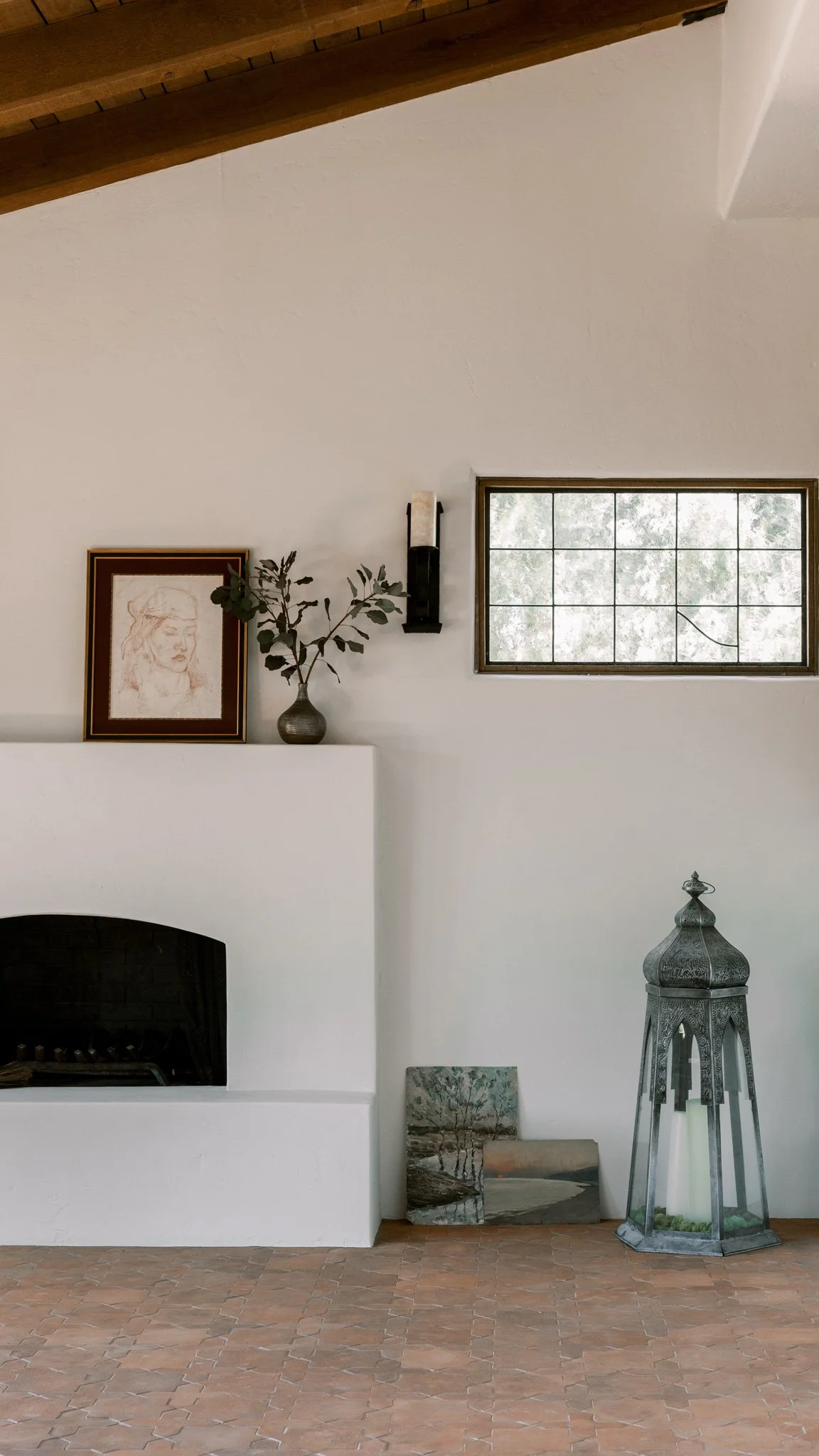 Interior living room corner with a white fireplace, framed sketch of a woman's face, a small vase with greenery, a wall sconce, two landscape paintings, a decorative lantern, and a small window.