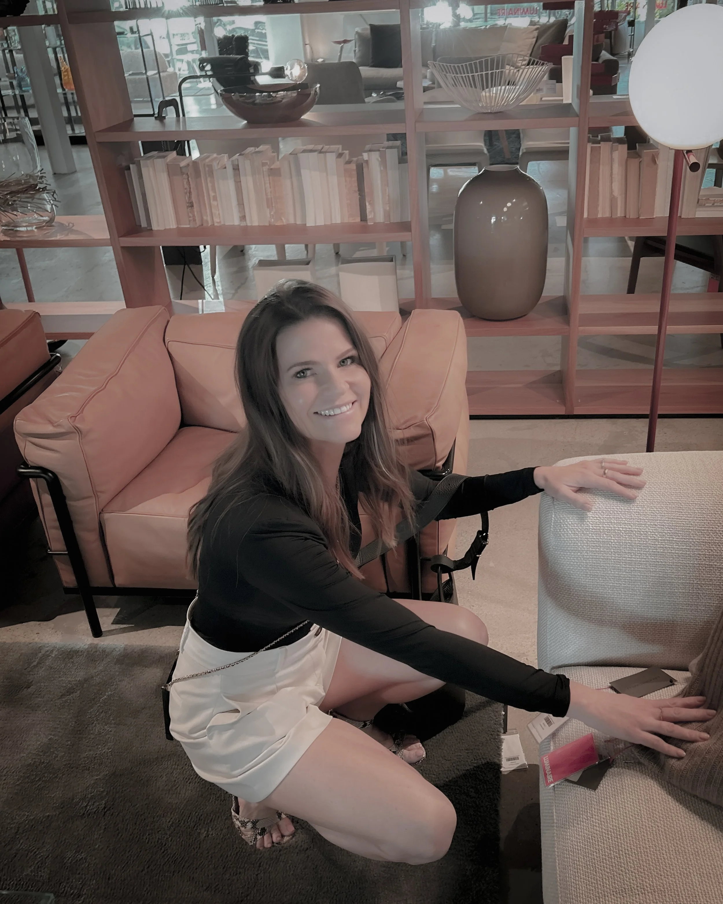 A woman with long brown hair smiling while crouching down in a furniture store, reaching for an item on a gray armchair. She is wearing a black top, white shorts, and sandals. The background shows a room divider with shelves and various decorative objects.
