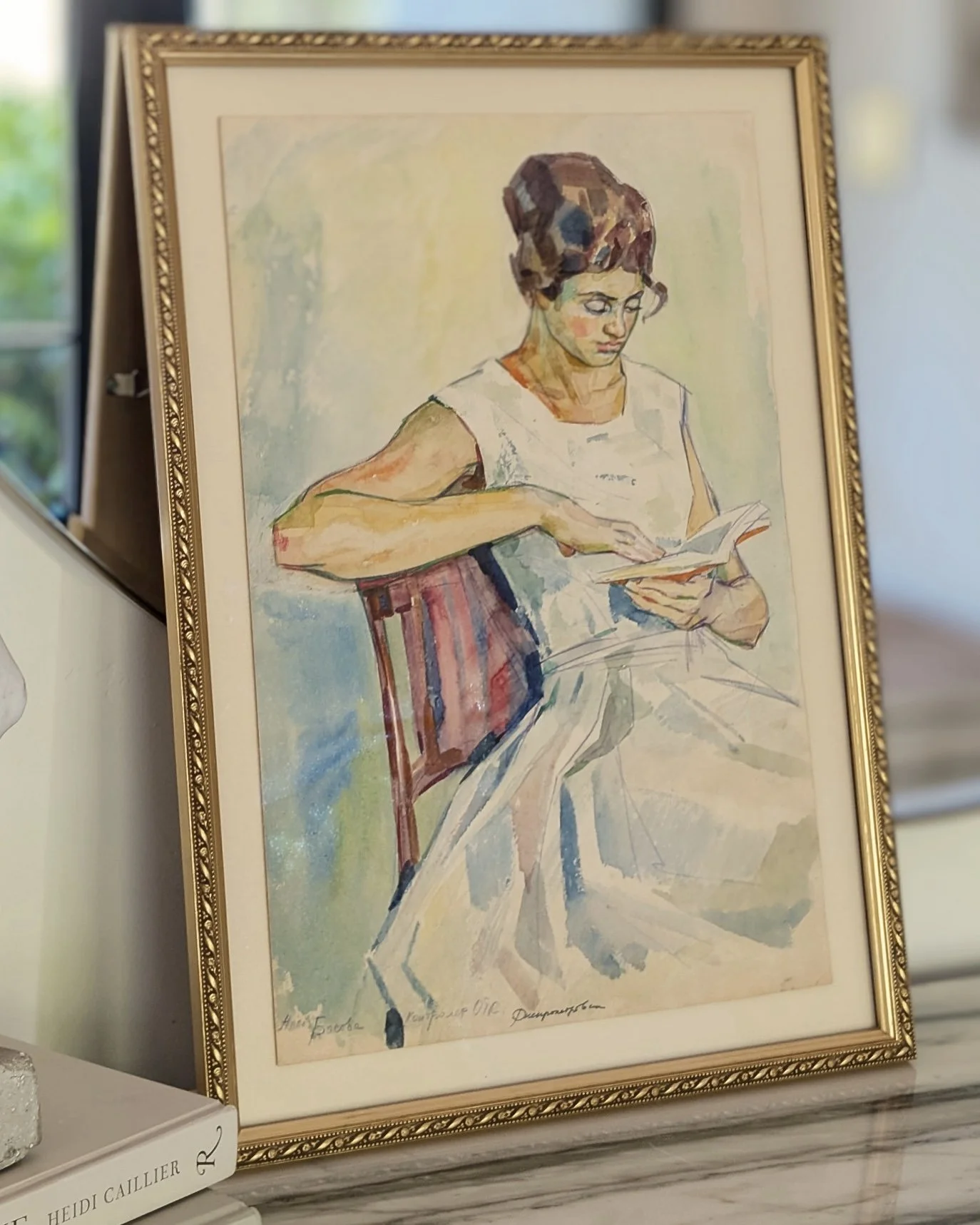 Watercolor painting of a woman reading a book, seated on a chair, with her hair in a bun, wearing glasses and a sleeveless white dress, framed in gold and displayed on a marble surface.