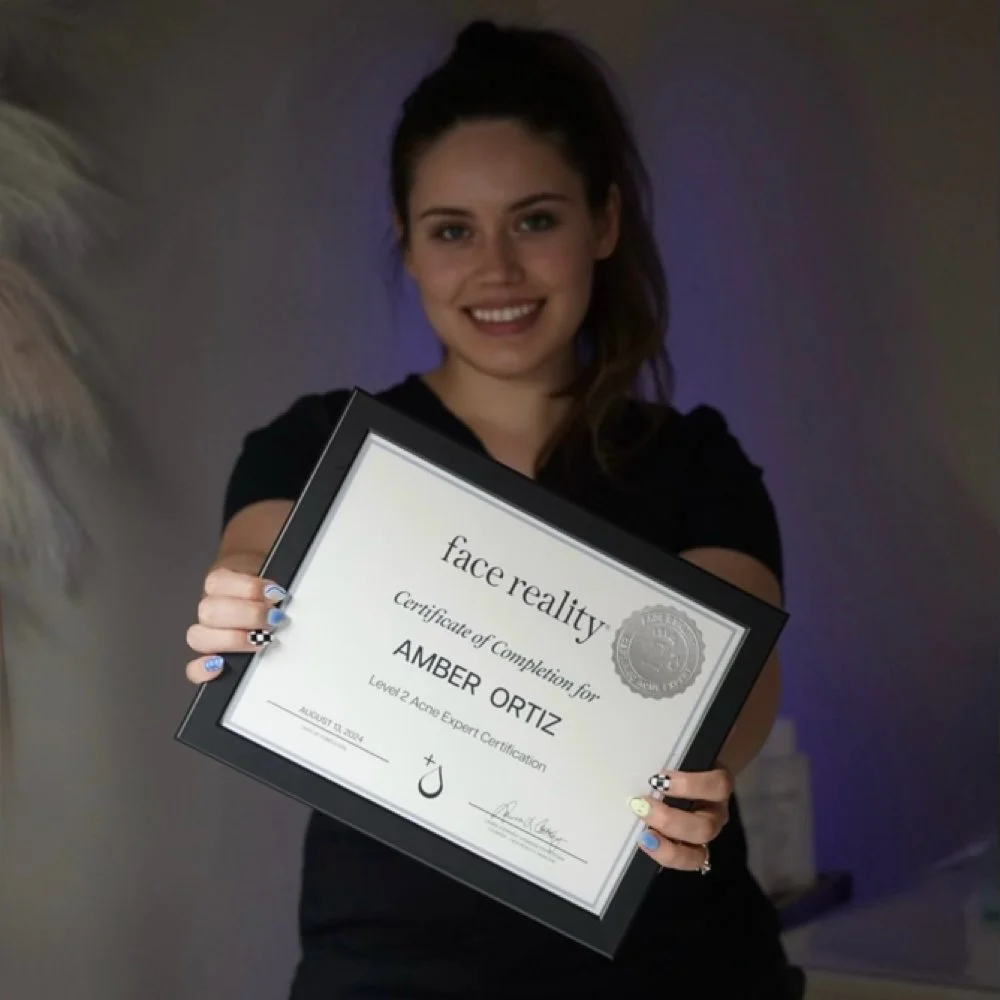 A young woman with long brown hair holding a certificate of completion for face reality. She is smiling and wearing a black shirt, with dark and colorful lighting in the background.