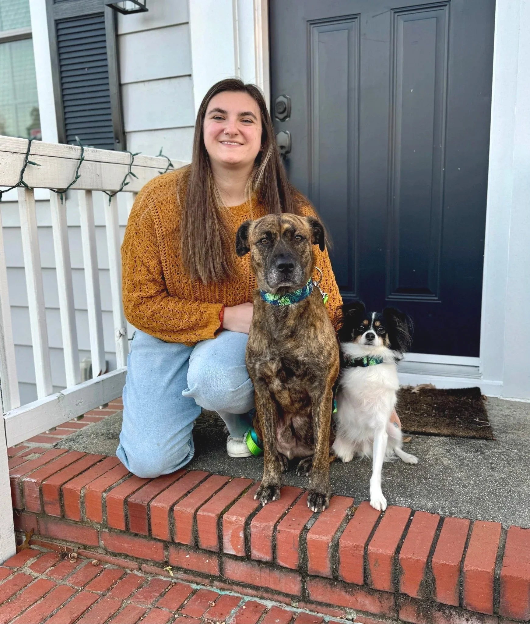 Photo of a female dog trainer in a mustard color sweater. To the right of the trainer is two dogs. One dog is brown and black. It's the same dog as the hero image. The other dog is on the far right. It's half the size of the brown and black dog.