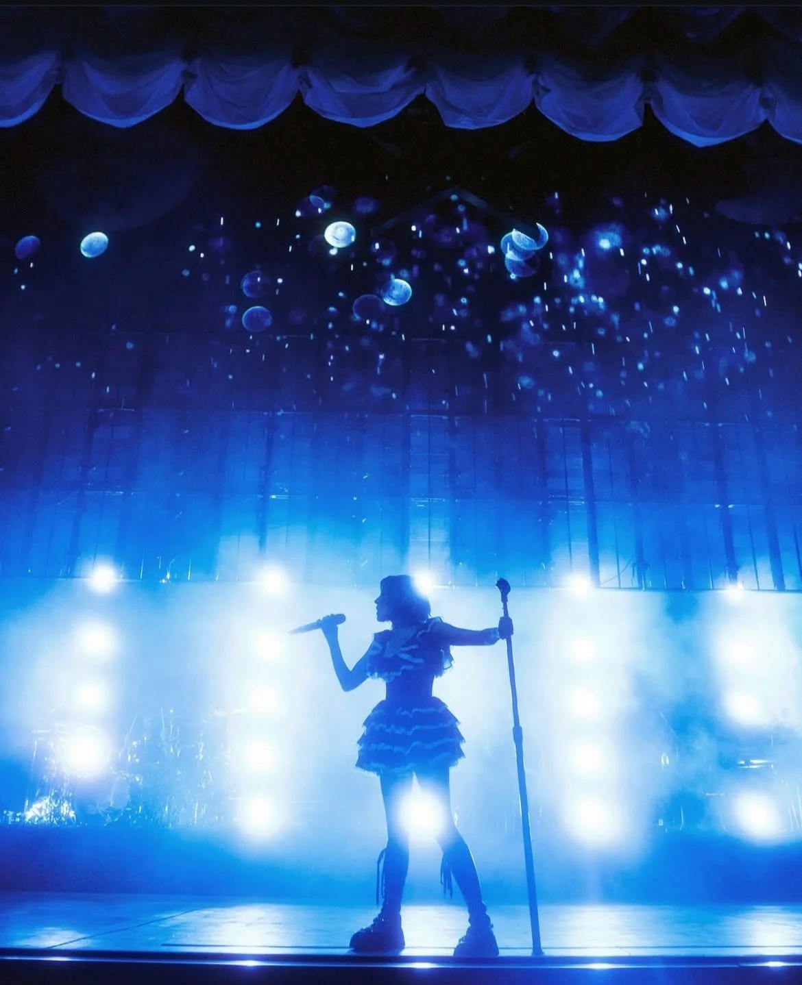 A silhouette of a female singer holding a microphone and a stand on stage with blue lighting and hanging decorations resembling jellyfish.