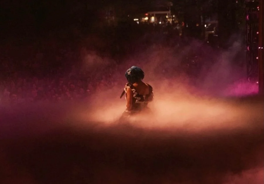 A person sitting on the stage in a dimly lit environment with pink and purple lighting, surrounded by smoke or fog.