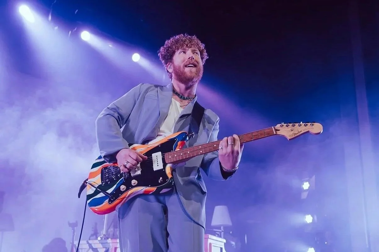 JP Saxe with curly hair and beard playing electric guitar on stage with blue and purple lighting.