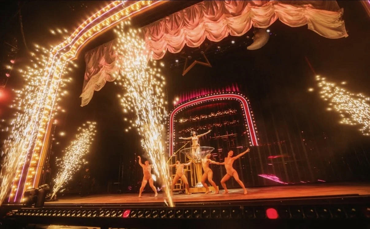 Circus performance with dancers, a central acrobat on a hoop, and fireworks on stage surrounded by pink drapes and star decorations.