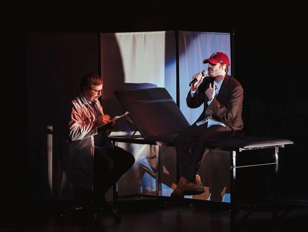 A stage scene with two men. One man is sitting on a chair, wearing a jacket and glasses, writing on a notepad. The other man, wearing a blazer, light shirt, and red cap, is sitting on a medical table, holding a microphone, and singing or speaking.