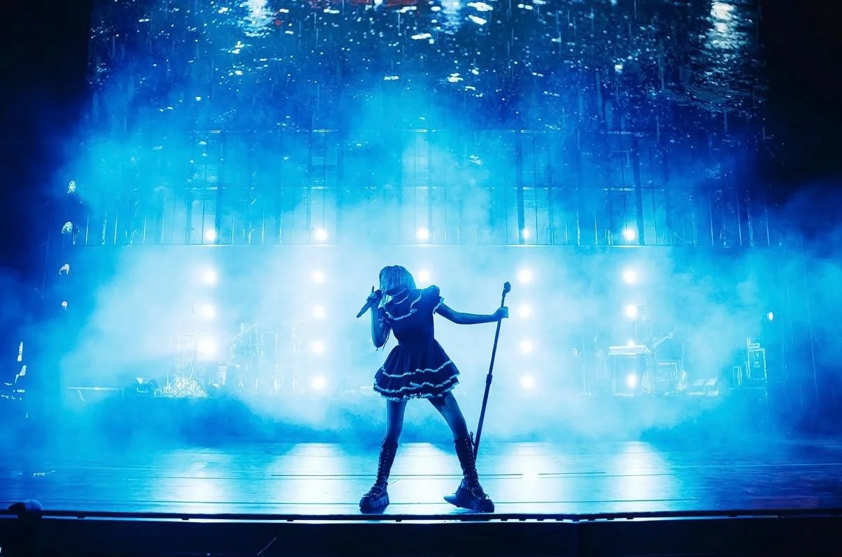 Performer singing on stage with a microphone, holding a staff, surrounded by blue stage lights and fog, with a reflective ceiling overhead.