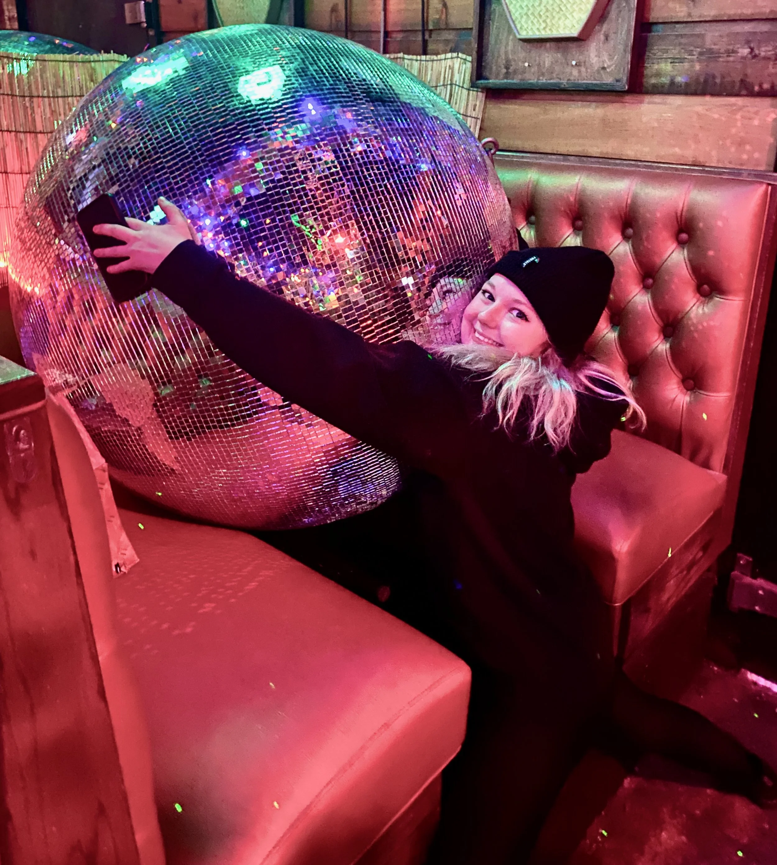 A woman smiling, wearing a black beanie and black jacket, takes a selfie with a large disco ball in a cozy, wood-paneled room with pink and red lighting.