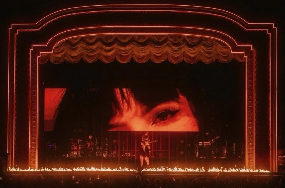 Stage with a large screen displaying a close-up of a woman's eye, illuminated by red lights, with a performer standing in front and musicians on the sides, and flames along the front of the stage.