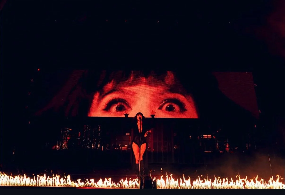 A stage with a performer in black costume and thigh-high boots, standing in front of a large screen displaying a close-up of a woman's eyes, with flames along the stage edge.