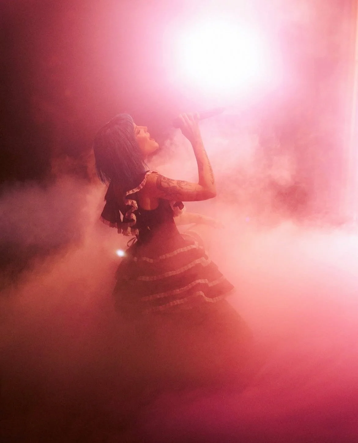 A woman with dark hair, tattoos on her arms, wearing a dress with ruffles, holding a microphone, standing in a smoky environment with pink and white lighting.