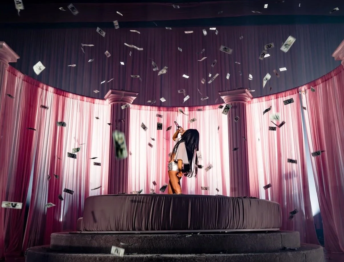A woman standing on a bed surrounded by falling dollar bills, singing into a microphone, with pink curtains in the background.