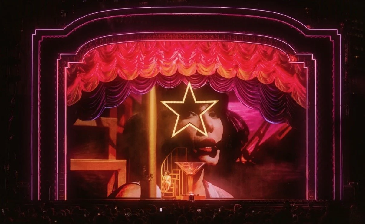 Stage with neon pink and purple curtains, a large star outline, and a background image of a woman's face with dark hair and makeup, in front of an audience.