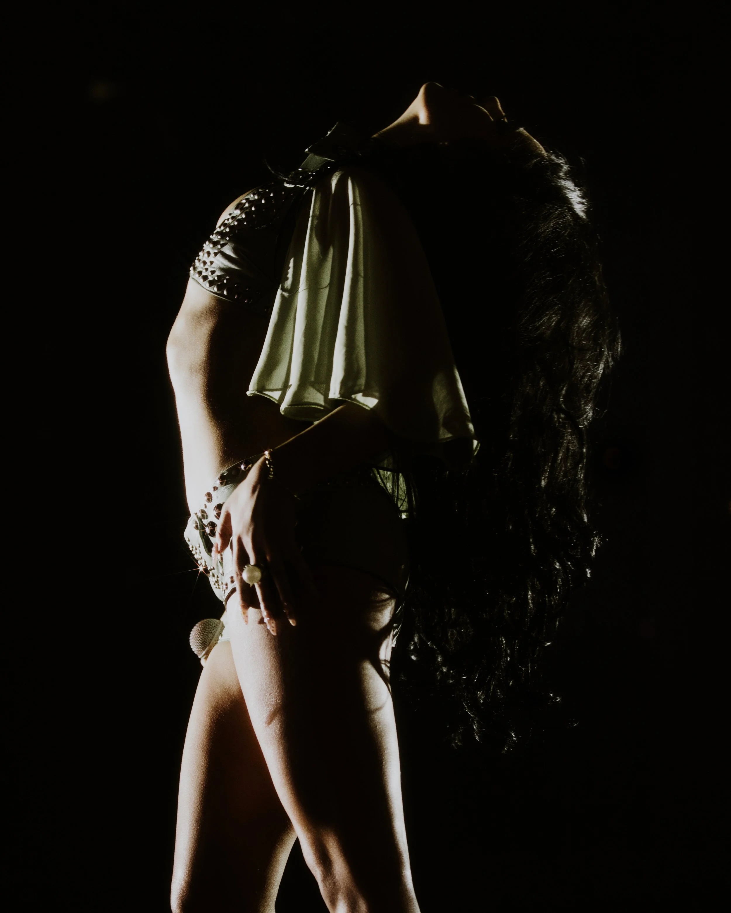 A woman with long curly hair, dressed in a shiny, studded top and skirt, is standing against a dark background, with her head tilted back and one hand raised near her face.