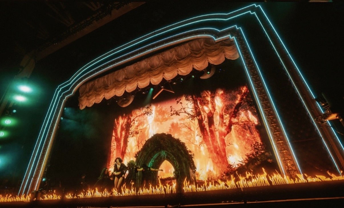A theatrical stage set depicting a fiery forest scene with flames and trees, illuminated by colorful lights and featuring actors performing.