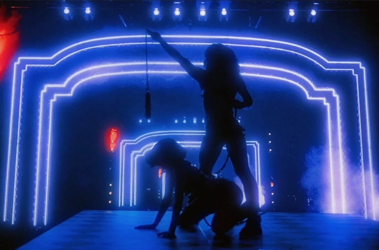 Two silhouetted performers on stage with neon blue and red lights in the background, one standing and pointing up, the other kneeling.