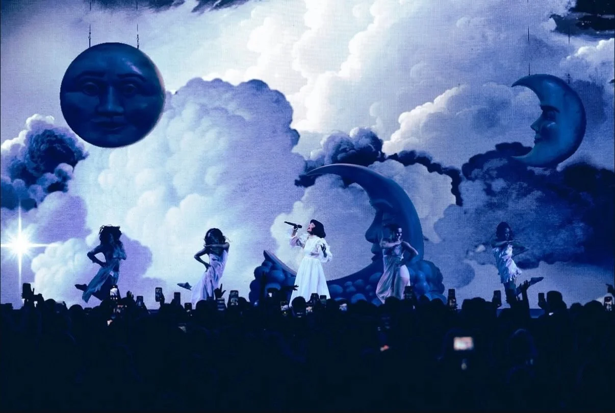 A stage performance with a surreal sky backdrop featuring large floating moon and sun faces, clouds, and a crescent moon with a face. Five women in white dresses are dancing, with one singing into a microphone. The audience is visible in silhouette, 