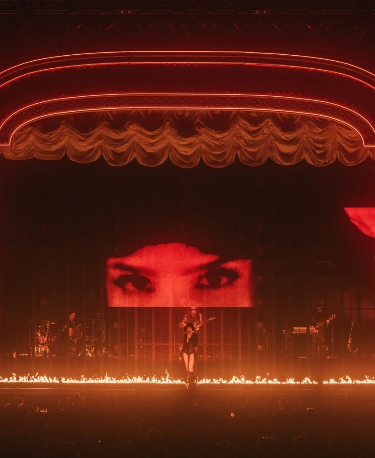 Stage with red lighting, a large screen displaying a close-up of eyes, a performer singing, band members playing instruments, and flames along the edge of the stage.