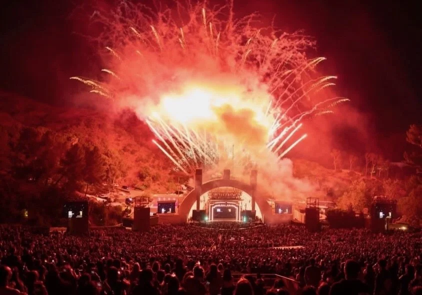 Large outdoor concert at night with fireworks illuminating the sky and a packed crowd watching.