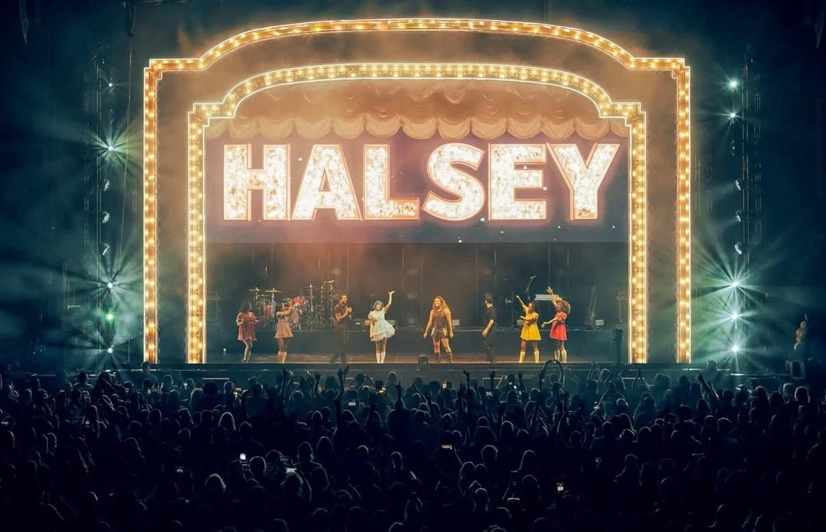 A concert stage with bright lights and a large illuminated sign reading 'HALSEY', with performers and dancers on stage and a crowd in front.