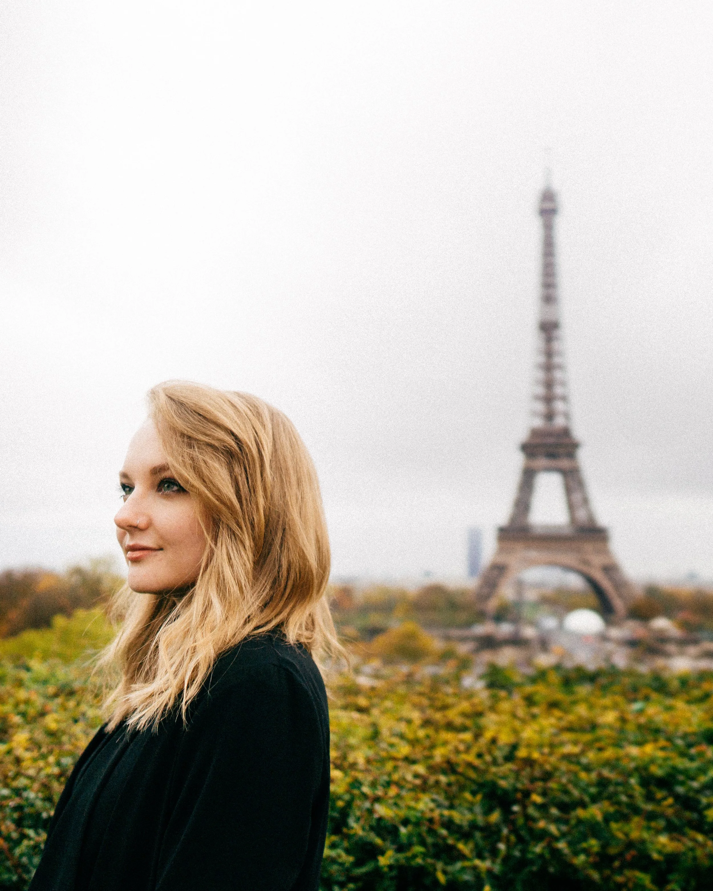 A woman with blonde hair and light skin standing outdoors, with the Eiffel Tower in the background on a cloudy day.