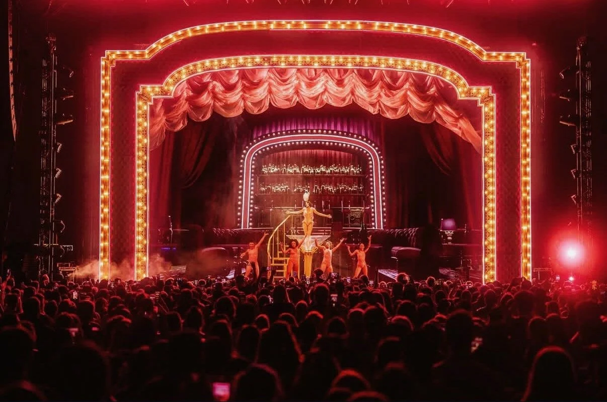 A theatrical stage with red curtains, illuminated with bright border lights, hosting a performance with a singer at center stage. Performers are dancing around her, with an audience watching.