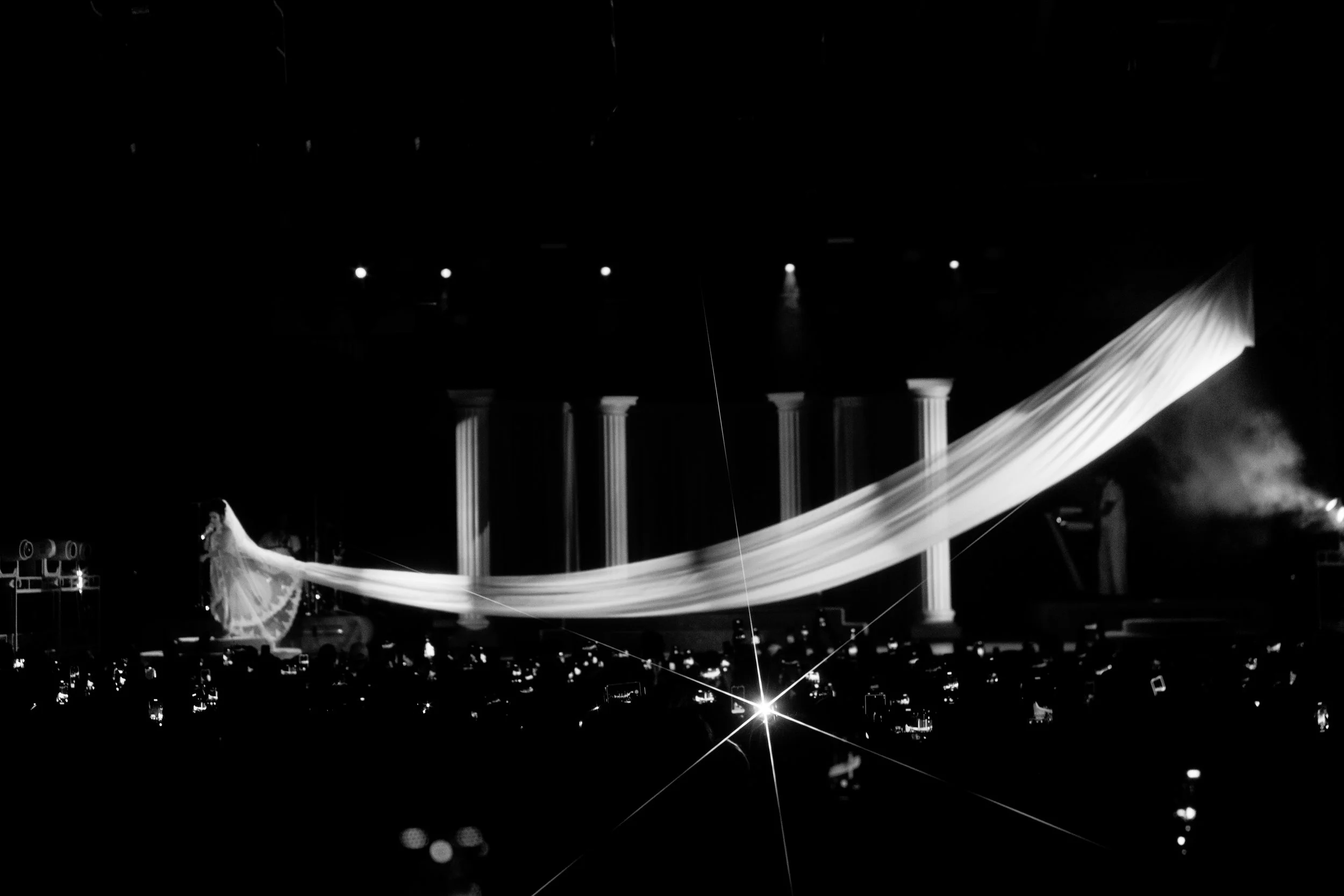 A black and white photo of a stage with columns and a flowing curtain. There is a performer dressed in white singing or speaking, with a microphone. The background has stage lights and equipment.