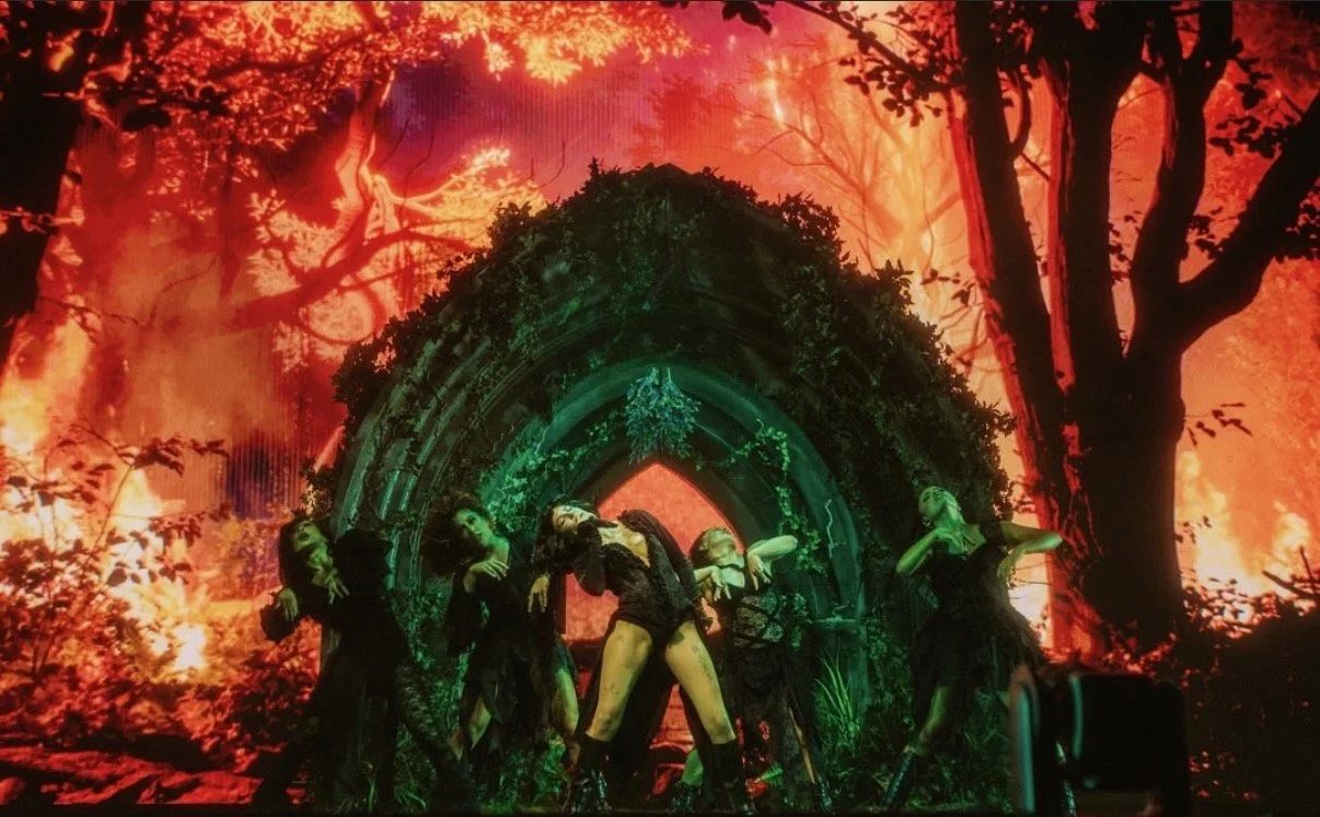 A group of five female dancers performs in front of a backdrop depicting a fiery forest with tall trees and flames. The dancers wear black outfits and strike dramatic poses near an arched stone structure covered in greenery.