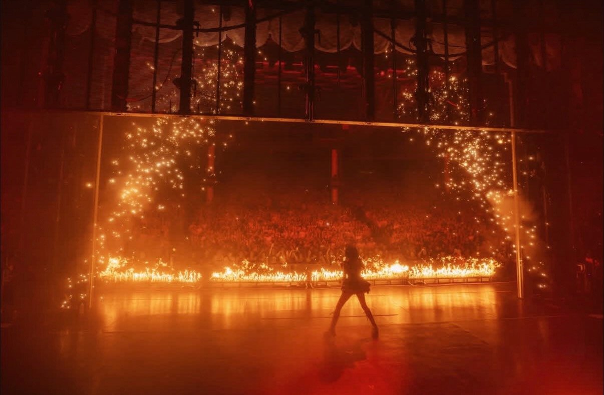 A person stands on a stage in front of a large, fiery spectacle with sparks and flames, possibly during a performance or event.