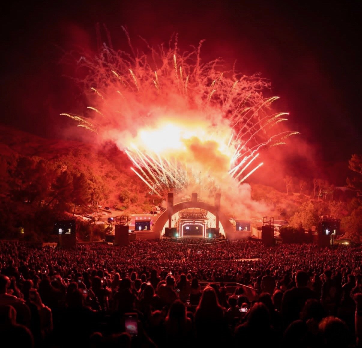 Large crowd watching fireworks display at night with a stage in front, set against a hillside.