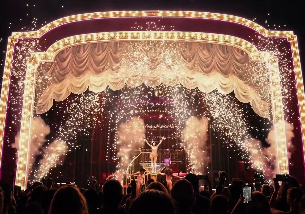 A circus or theatrical stage with performers and a person standing in the center with arms raised, surrounded by sparks and smoke, with an ornate, illuminated curtain at the top.