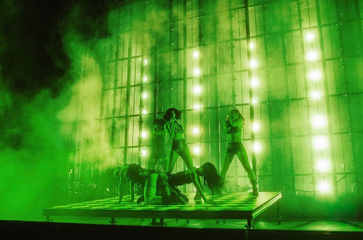 Five performers on a checkered stage, with three in a crawling position and two standing, surrounded by green fog and bright lights forming a backdrop.