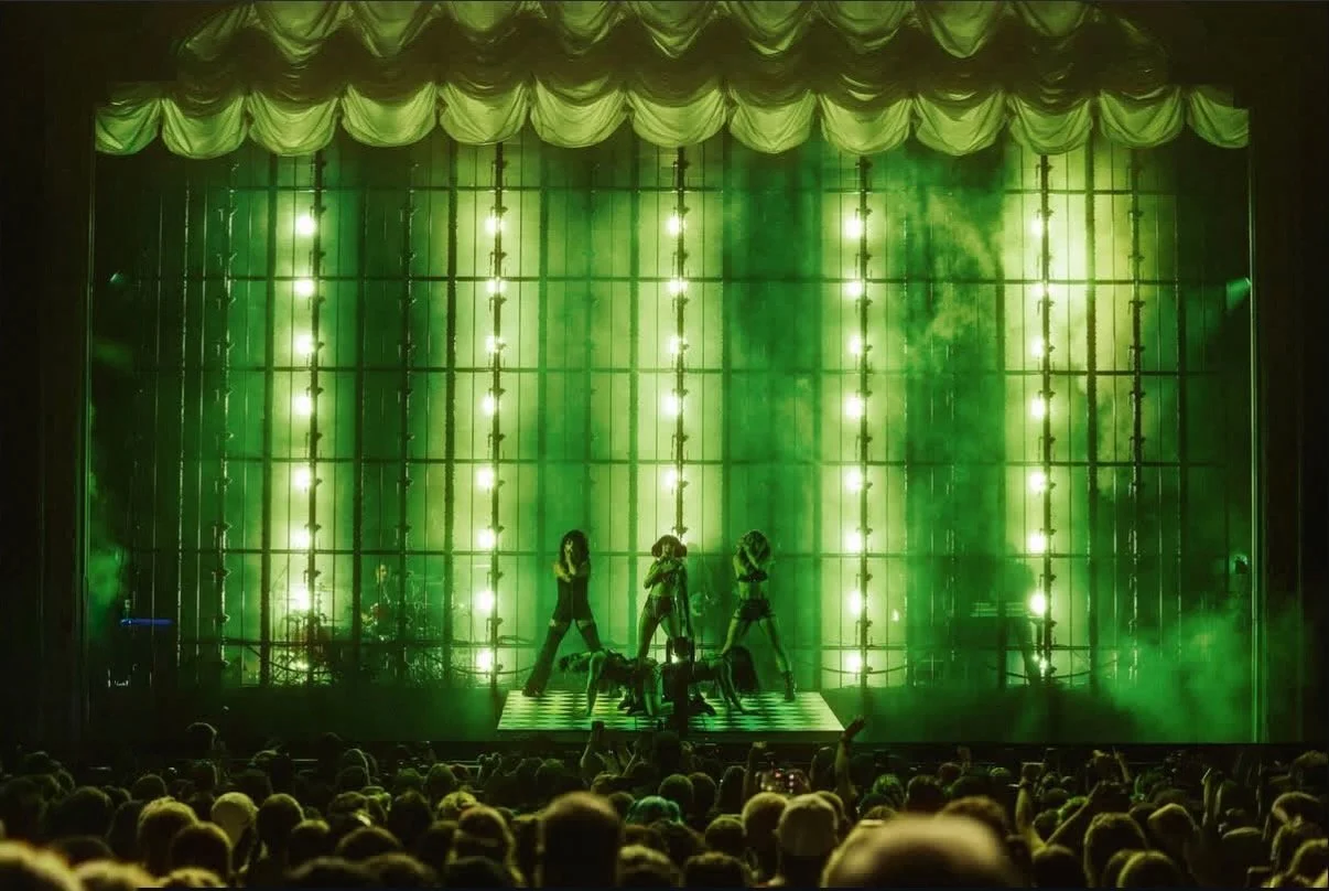 Performers on stage with green lighting and fog effects, audience watching in the foreground.