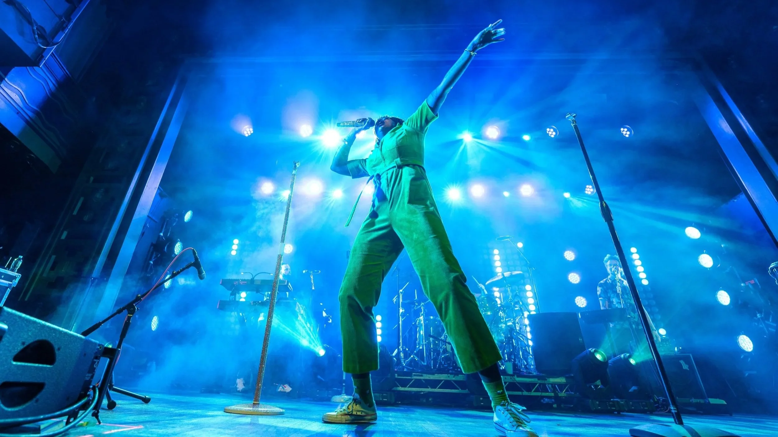 A performer on stage singing into a microphone, with one arm raised and surrounded by bright blue stage lights, music equipment, and band members in the background.