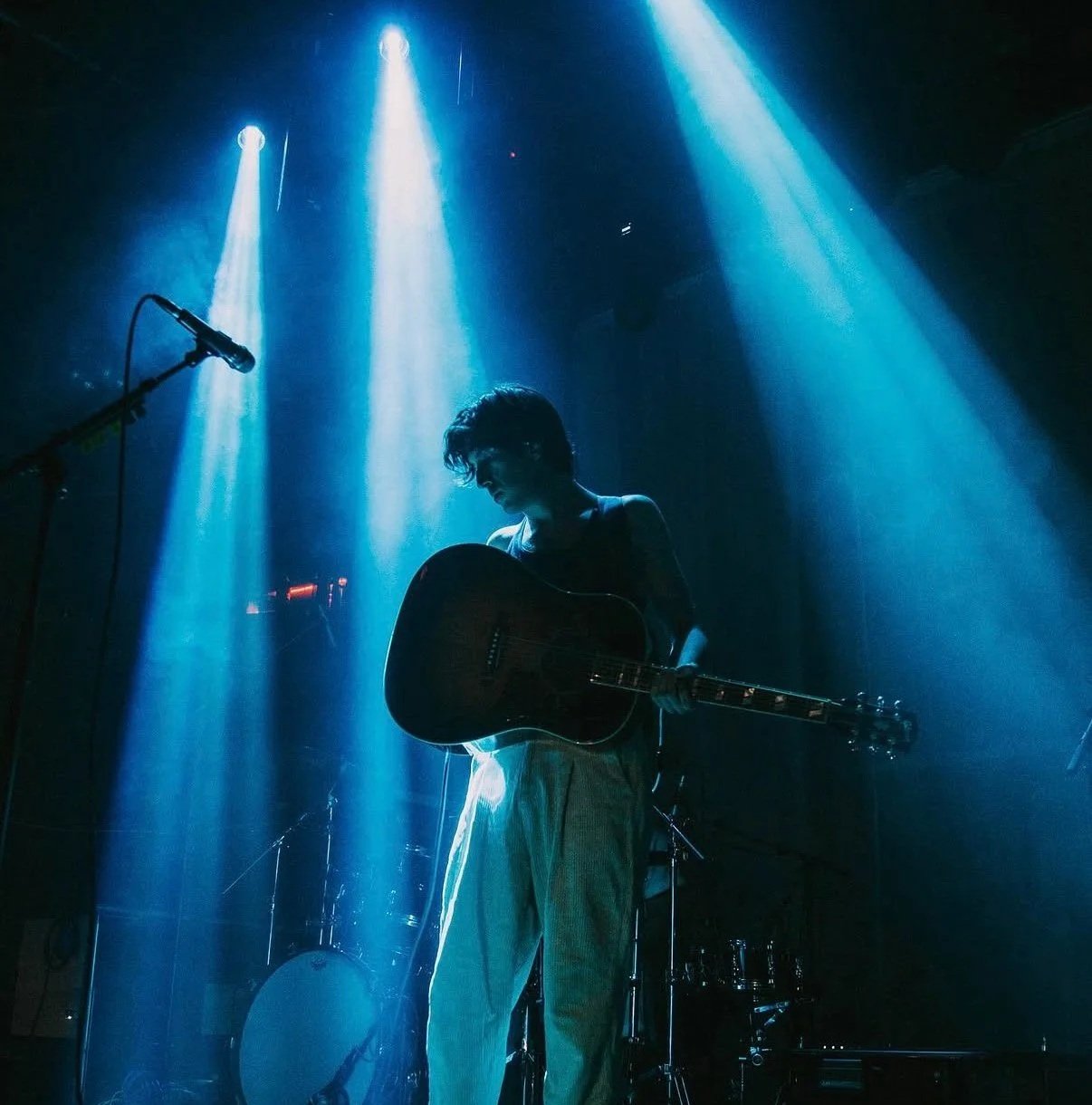 A person with short curly hair playing an acoustic guitar on stage, illuminated by blue stage lights, with a microphone stand nearby, and drums in the background.