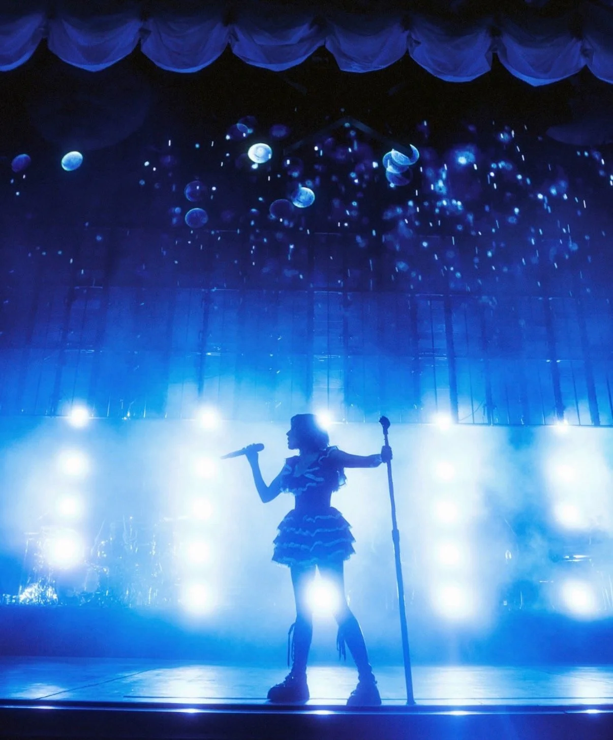 A silhouette of a performer on stage holding a microphone in one hand and a staff in the other, with bright blue stage lighting and a backdrop of hanging blue fabric and floating jellyfish-like objects.