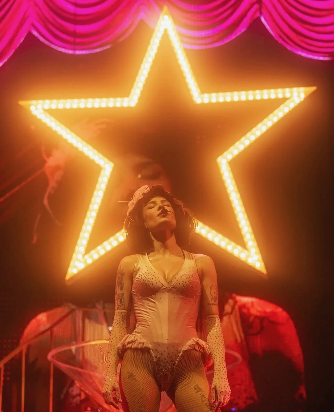 A womanm, Halsey, standing in front of a large illuminated star on a pink curtain backdrop, dressed in a revealing outfit with tattoos visible on her arms, with a confident expression and closed eyes.