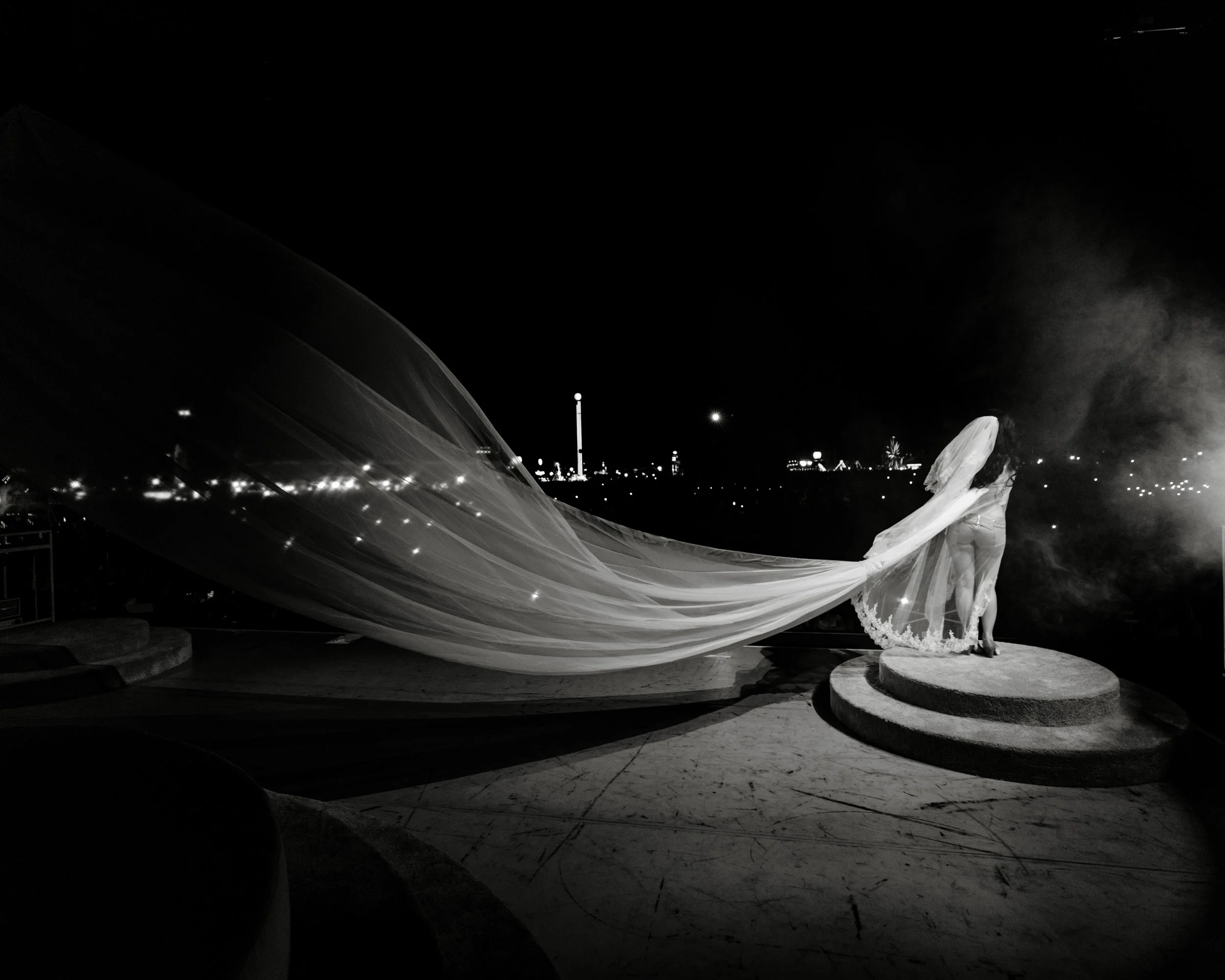 Black and white photo of a statue of a woman with a long flowing dress, standing on a circular base outdoors at night, with city lights, a Ferris wheel, and a tall building illuminated in the background.