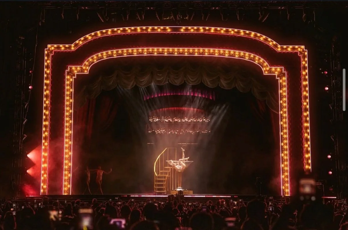 Theater stage with bright lights and red velvet curtains, featuring a performer on stilts and a cheerleader in the background, with an audience in front.