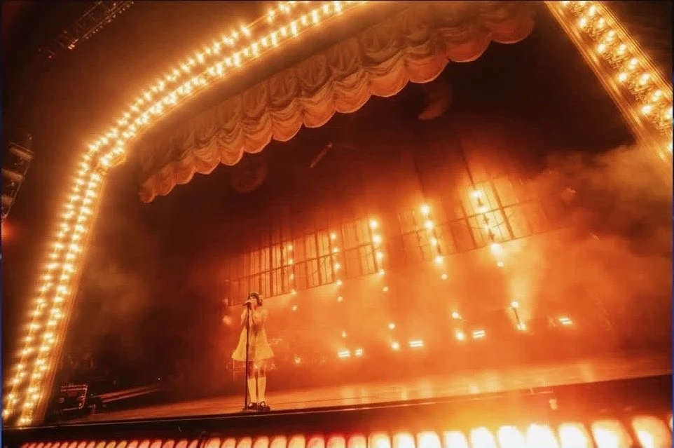 A performer standing on a stage with bright lights and a fiery orange glow, with curtains hanging above, creating a dramatic theater atmosphere.