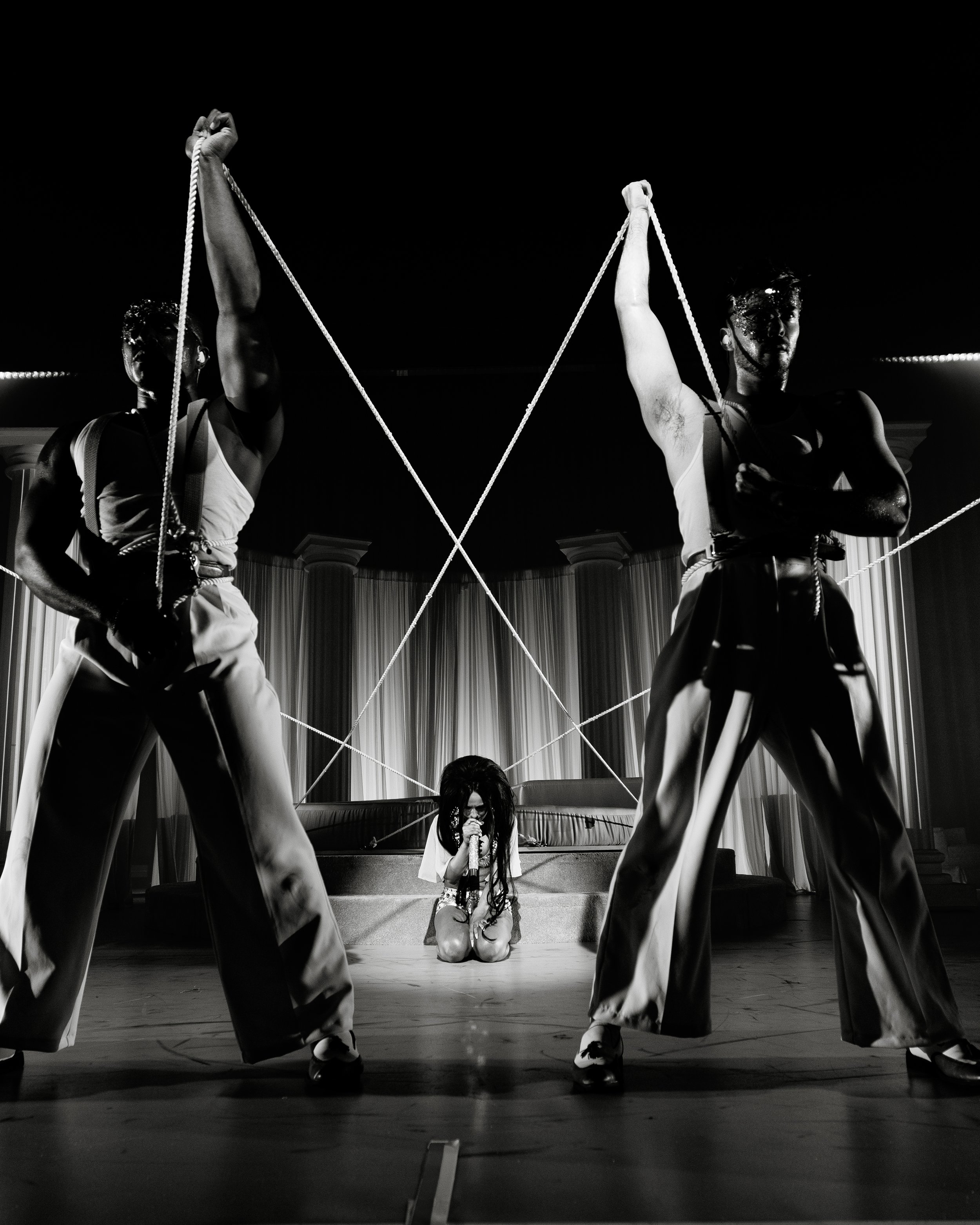 Black and white photo of a performer with long hair, sitting on the floor and singing into a microphone, with two dancers standing behind them, wearing matching outfits and holding ropes tied to their wrists, in a theatrical stage setting.