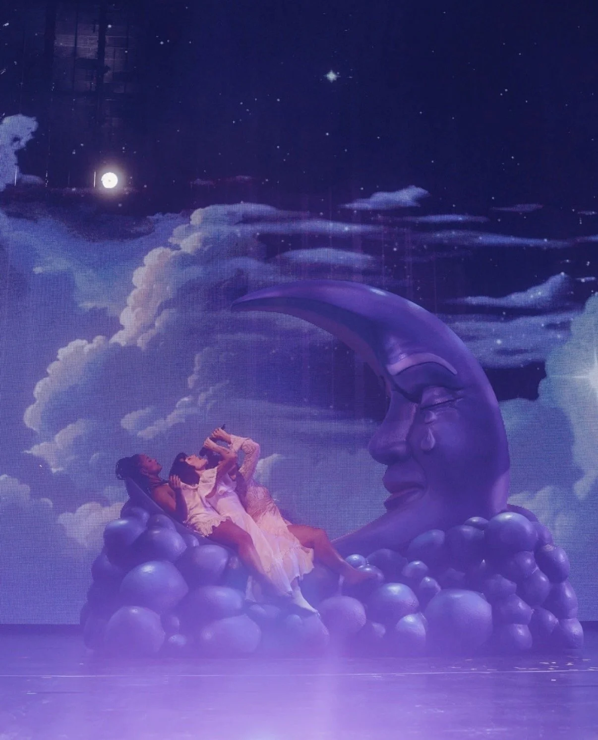 Three children in white dresses sitting on a cloud-shaped platform in front of a large moon sculpture with a human face, set against a background of a starry night sky and clouds.