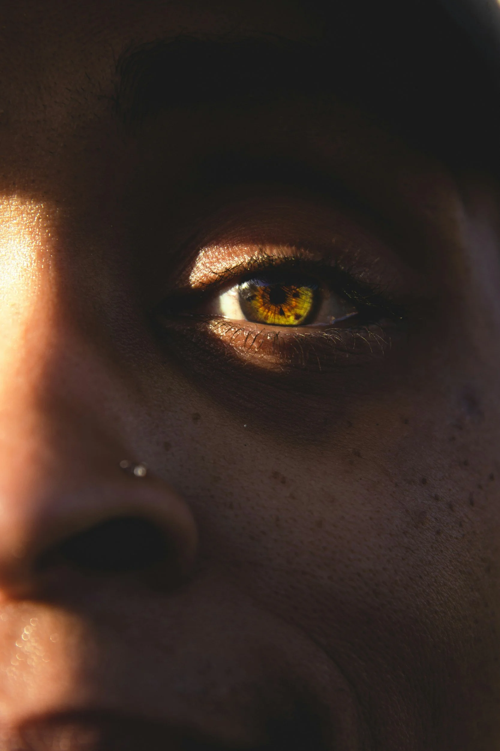 Close-up of a person's face showing an eye with bright hazel iris, illuminated by sunlight, with skin texture and makeup visible.