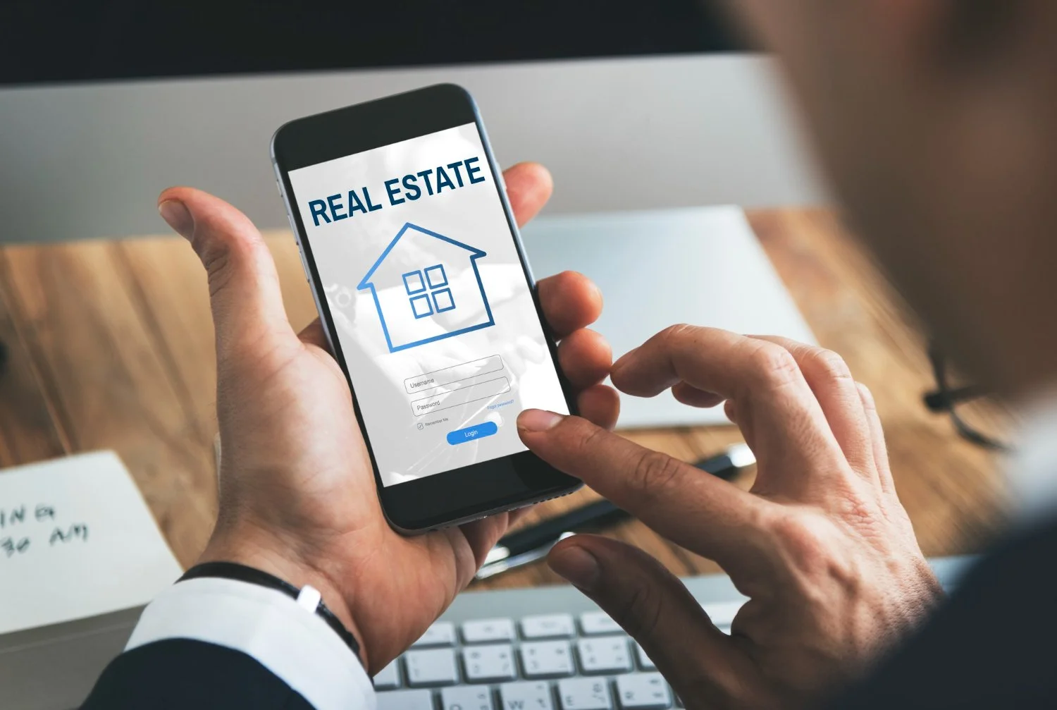 Person holding a smartphone displaying a real estate login screen with house icon, username, password fields, and a blue login button.