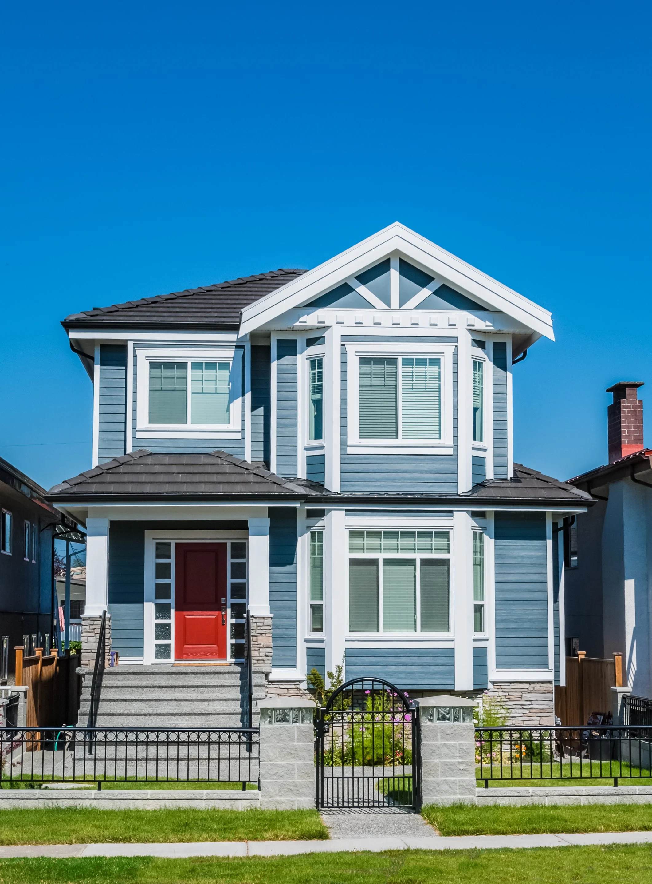 A two-story blue house with a red front door, large windows, white trim, a small front yard with grass, and a black metal fence and gate under a clear blue sky.